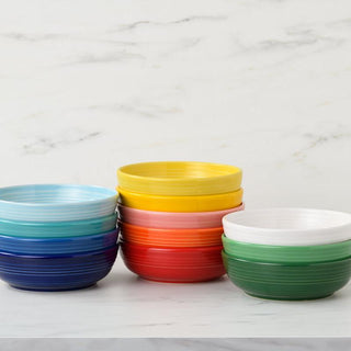 A stack of colorful Fiesta Coupe 6 3/4 Inch Salad Bowls (24 OZ) in blue, yellow, red, white, and green are displayed in two piles on a white marble surface against a light background.