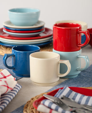 A set of Fiesta 16 OZ Stackable Mugs in red, blue, cream, and light blue sits with matching plates and bowls on a table—ideal for a coffee bar setup—with napkins and utensils arranged nearby.