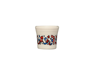 The Fiesta 4th of July 1.5 oz ceramic shot glass, made in the USA, features a cream color with red, white, and blue fireworks, rockets, stars, and streamers decorating the rim against a plain white background.