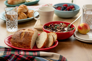 A breakfast table set with sliced bread on the Fiesta 2-Piece Serve Set 11 5/8 Inch Oval Platter & 38 OZ Bistro Bowl, a bowl of salad, an apple, croissants, cups, water glasses, berries in a Bistro bowl, and a plaid cloth in the foreground.