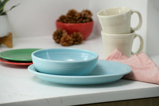 A Fiesta 2-Piece Serve Set—an 11 5/8-inch oval platter and a 38 oz bistro bowl—sits on a white countertop next to a pink cloth, with cream mugs, colorful plates, and pine cones in a red bowl in the background.