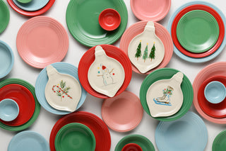 A collection of ceramic plates and bowls in festive holiday colors, including red, green, pink, and light blue, with some featuring hand-painted winter illustrations like snowmen, Christmas trees, and candy canes.