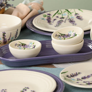 A set from the Lavender Bloom Collection features ceramic dishes adorned with lavender flower designs—plates, bowls, and small dishes—arranged on a table with a purple tray, napkins, and a bundle of dried lavender. The ensemble includes the Fiesta Lavender Bloom 3 3/4 Inch Mini Bowl 5 OZ for added charm and versatility.