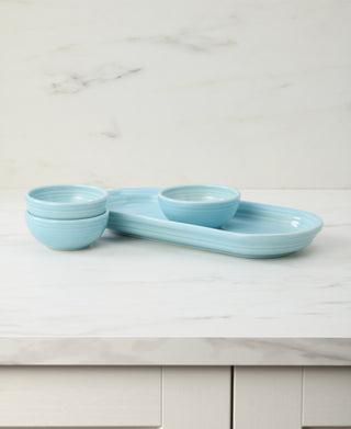 The Fiesta 4-Piece Condiment Set Sky by Fiesta features a light blue serving tray with two matching mini bowls on top and two more stacked nearby, displayed on a white marble countertop with a marble-patterned background.