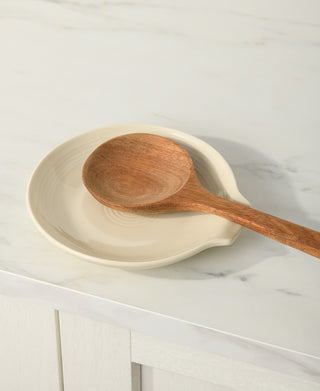 A wooden spoon rests on the Fiesta Extra Large Spoon Rest 7 Inch, placed on a round cream-colored plate atop a white marble countertop.