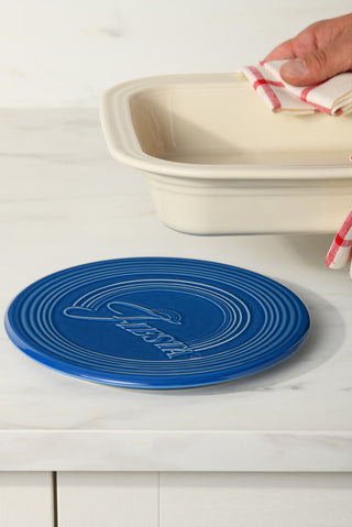 A hand with a red-and-white striped towel holds a cream dish above the Fiesta Extra Large Trivet 8 1/4 Inch, a heat-resistant ceramic trivet embossed with "Fiesta," on a white marble countertop.