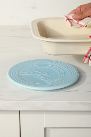 A Fiesta Extra Large Trivet 8 1/4 Inch in light blue rests on a white countertop. In the background, a hand with a red-and-white towel holds a beige ceramic baking dish.