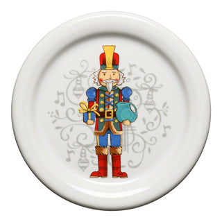 The Fiesta Nutcracker Coaster Set of 2 features a decorative white surface with an illustrated nutcracker holding a gift and blue ornament, plus festive patterns and musical notes in the background.