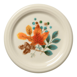 The Fiesta Autumn Fields 4 3/8 Inch Coaster and Mug Cover is a round, cream-colored item made in the USA, decorated with orange and yellow autumn leaves, blue-green leaves, brown acorns, and clusters of blue dots.