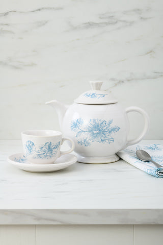 A Fiesta Retired Botanical Floral Classic Rim 5 7/8 Inch Saucer sits on a white marble countertop with a white teapot and teacup featuring blue floral designs, plus a folded matching napkin and a spoon.