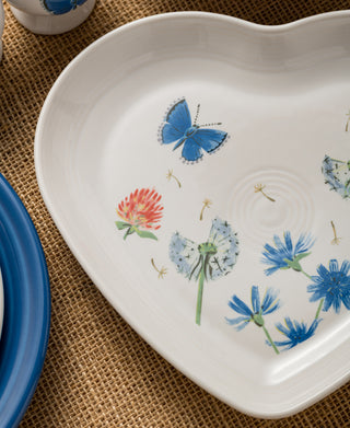 The Retired Breezy Floral 9 Inch Heart Shaped Plate by Fiesta, made in the USA, features blue butterflies, dandelions, red clover, and wildflowers. Displayed on woven burlap beside blue-rimmed dishes, it adds charm to any table.