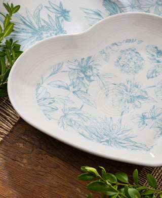 The Retired Fiesta Botanical Floral 9 Inch Heart Shaped Plate by Fiesta features blue floral designs and is displayed on a rustic wooden table with green leaves and a matching floral cloth in the background.