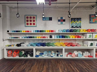 Shelves display colorful plates, bowls, and mugs arranged by color in a bright, modern room with white brick walls, wooden floor, and framed art hanging above the shelves.