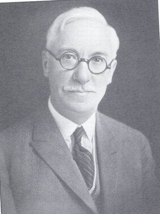 Black and white portrait of an older man with glasses, white hair, and a mustache. He is wearing a suit, tie, and vest, and is looking directly at the camera with a neutral expression.