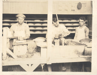 Three men working in a bakery; one kneads dough at a counter, while the other two prepare baked goods, surrounded by trays and supplies, with shelves of bread in the background.
