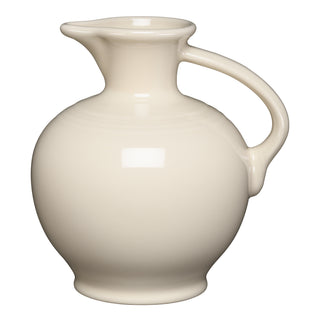 The Fiesta Linen Carafe is a cream-colored ceramic pitcher with a round body, narrow neck, curved spout, and large handle. Made in the USA from lead-safe china. Shown against a plain white background.