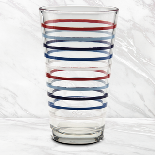 The 16 oz. Fiesta Americana Thin Stripe Cooler Glass by Moderne Glass features horizontal red, blue, and light blue stripes and sits on a white marble surface with a marble background. Sold in sets of 4.