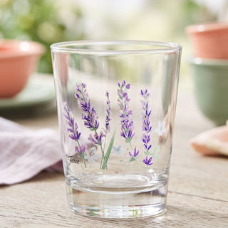 A set of four Moderne Glass Fiesta Lavender Bloom 15 oz. Tapered Double Old Fashion glasses, made in the USA and adorned with painted lavender flowers, sits on a wooden table with blurred Fiesta Dinnerware plates, bowls, and greenery in the background.