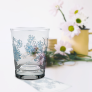 A Moderne Glass Botanical Floral 15 oz. Tapered Double Old Fashion Glass from a set of 4, decorated with light blue flowers, rests on a white surface. In the softly blurred background is a Fiesta Dinnerware yellow pitcher with pink and white daisies.