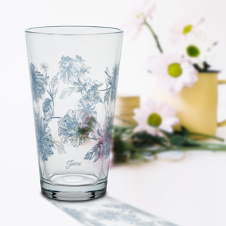 A Moderne Glass Botanical Floral 16 oz. Cooler Glass, decorated with blue floral designs, stands on a white surface. In the background, pink daisies fill a yellow Fiesta Dinnerware watering can vase.