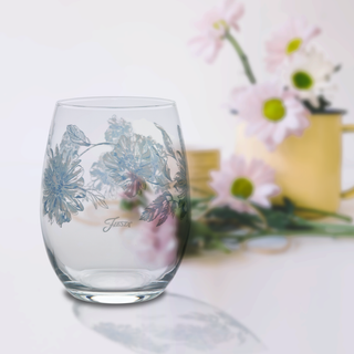 The Moderne Glass Botanical Floral 15 oz. Stemless Wine Glass set of 4 features a delicate floral design on clear glass. Made in the USA, these glasses bring botanical charm to your table. Light pink and white daisies and a yellow watering can create a blurred backdrop.