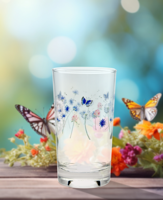 A set of four Retired Breezy Floral 7 oz. Juice Glasses by Moderne Glass, decorated with blue flowers and butterflies, is displayed on a wooden surface among colorful blossoms against a softly blurred natural backdrop.