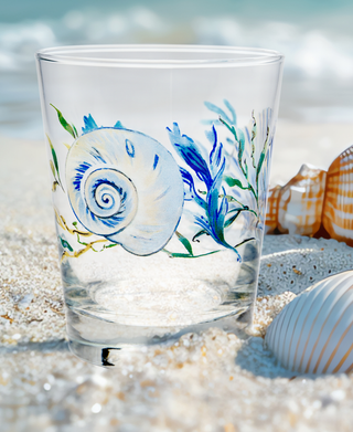 A set of four Retired Coastal Shores 15 oz. Tapered Double Old Fashion Glasses by Moderne Glass, featuring a blue and white seashell design, rests on a sandy beach with real shells and ocean waves in the background.