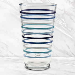A 16 oz. Fiesta Coastal Blues Thin Stripe Cooler Glass by Moderne Glass, featuring horizontal blue and navy stripes, sits on white marble with a matching background. "Jassni" is printed near the bottom. Set includes 4 glasses.