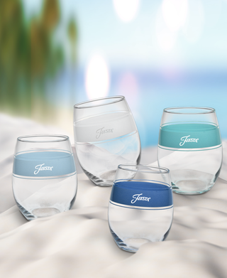 A set of four Moderne Glass Coastal Blues 15 oz. Frame Stemless Wine Glasses in mixed blue and clear bands sit on a sandy beach with palm trees and ocean in the background, evoking a relaxed, tropical vibe. Made in the USA.