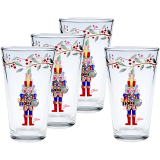The Moderne Glass Nutcracker The Giver 16 oz. Cooler Glass Set includes four glasses with painted nutcracker soldiers and a festive holly berry garland, perfect for Christmas celebrations. Designs face forward and glasses are arranged in two rows.