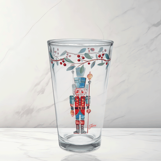 The Moderne Glass Nutcracker The Guardian 16 oz. Cooler Glass Set includes 4 clear glasses featuring a festive nutcracker in red, blue, and green with a leaf and berry garland—ideal for holiday gatherings. Shown on white marble background.