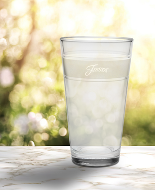 A Linen 16 oz. Frame Cooler Glass by Moderne Glass sits on a marble surface with "Fiesta" etched on it, sunlight and greenery blurred in the background—ideal for Fiesta Dinnerware fans. Set includes 4 glasses.