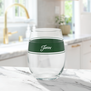 A 15 oz. Fiesta® Frame Stemless Wine Jade glass by Moderne Glass, featuring a green band and "Fiesta" imprint, sits on a white marble counter with a blurred sink and faucet in the background. Set of 4, made in the USA.
