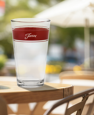 A set of 16 oz. Fiesta® Frame Coolers in Scarlet by Moderne Glass sits on an outdoor wooden table with blurred chairs and a white umbrella in the sunny background. Made in the USA.