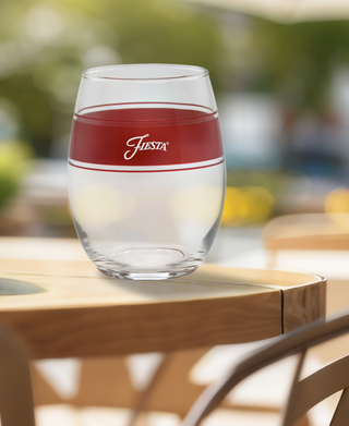 A set of four 15 oz. Fiesta® Frame Stemless Wine Scarlet glasses by Moderne Glass sits on a wooden outdoor table with greenery and yellow flowers blurred in the background. Made in the USA.