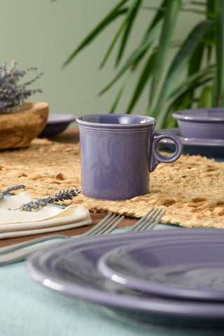 A Fiesta Lavender Classic Ring Handle 10 1/4 OZ Mug rests on a woven placemat with matching dinnerware and cutlery, accented by a sprig of lavender and greenery for a cozy, stylish table setting.