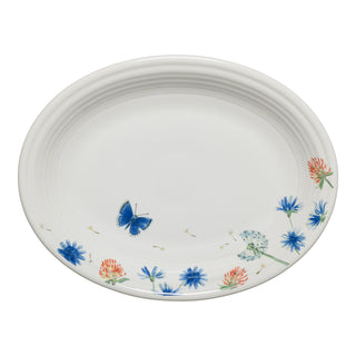 The Fiesta Retired Breezy Floral 11 5/8 Inch Medium Oval Serving Platter, made in the USA, features a white ceramic base with blue and red wildflowers, green leaves, a dandelion, and a blue butterfly near the edge.