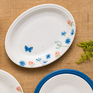 The Fiesta Retired Breezy Floral 11 5/8 Inch Medium Oval Serving Platter in white vitrified china features blue and coral flowers, a butterfly, and green leaves along the rim, placed on burlap beside another plate with a blue rim and yellow details.