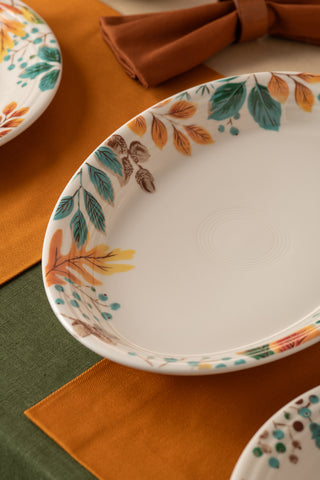 The Fiesta Autumn Fields 13 5/8 Inch Large Oval Serving Platter, made in the USA from durable vitrified china, features colorful autumn leaf patterns and is shown on an orange and green tablecloth with a brown napkin.