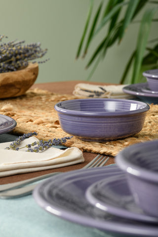 A close-up of a table set with Fiesta Lavender Classic Rim 5 5/8 Inch Small Bowl, matching plates, cream napkins, silver cutlery, a woven placemat, sprigs of lavender, and in the background, a green plant and wooden bowl of lavender.