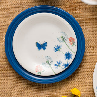 The Fiesta Retired Breezy Floral Classic Rim 7 1/4 Inch Salad Plate features blue edges, floral designs, and a blue butterfly on white vitrified china. Shown on a burlap tablecloth with another plate and yellow flowers nearby. Dishwasher safe.