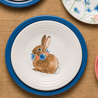 The Fiesta Retired Breezy Floral Easter Classic Rim 9-inch Luncheon Plate, made in the USA, features a brown rabbit with a flower wreath and a blue rim. It rests on burlap with other floral plates visible nearby.