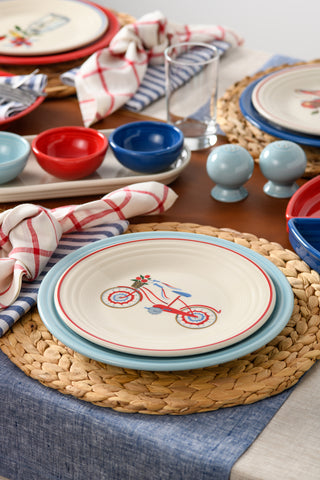 A close-up of a dining table features woven placemats, blue and red dishes, colorful bowls, striped napkins, and the Fiesta Americana Bicycle Classic Rim 9 Inch Luncheon Plate for a casual yet festive patriotic setup.