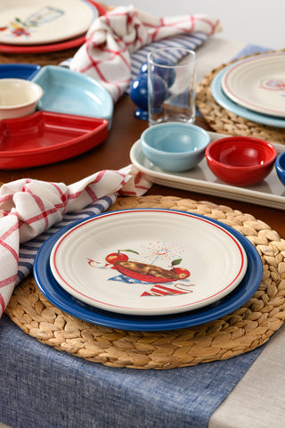 A festive table setting showcases the Fiesta Americana Apple Pie Classic Rim 9 Inch Luncheon Plate, paired with a woven placemat, colorful bowls, red and blue plates, a glass, and a white napkin with red checks for summer picnic flair.