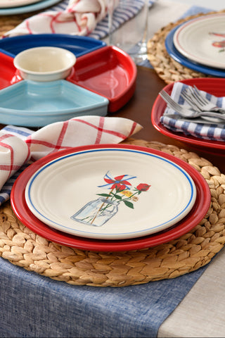 A close-up of a table set with the Fiesta Americana Mason Jar Classic Rim 9 Inch Luncheon Plate, red charger, woven placemat, checkered napkins, and divided serving dishes in patriotic colors for a festive look.