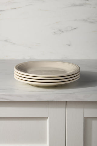 A set of four Fiesta Linen Classic Rim 10 1/2 Inch Dinner Plates is stacked on a marble countertop with a white and gray marble backsplash and white paneled cabinets below.