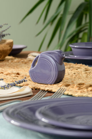 The Fiesta Lavender 5 OZ Mini Disk Pitcher rests on a woven placemat, paired with lavender plates, silverware, and greenery in the background for a cozy and elegant table setting.