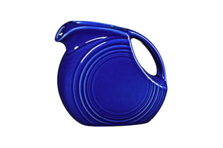 The Fiesta Retired Small Disk Pitcher is a round, cobalt blue ceramic pitcher made in the USA with an Art Deco design—concentric circles and a curved handle—shown on a white background.