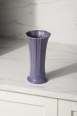 The Fiesta Lavender 9 5/8 Inch Medium Vase sits atop a white marble countertop, its tall fluted design and scalloped rim set against a light-colored marble backsplash.