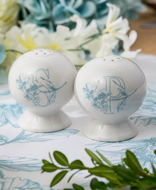The Fiesta Botanical Floral Sky 2 5/8 Inch Salt and Pepper Shaker Set features white ceramic shakers with floral designs and blue S and P letters, displayed on a patterned tablecloth amid green leaves and white flowers.